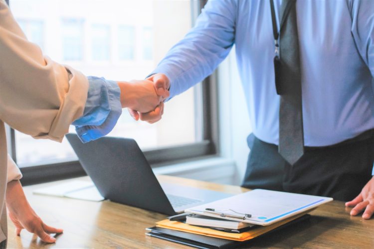 Two business men shaking hands over a desk with a laptop and files