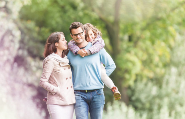 A young family walk through the park with the dad carrying the daughter