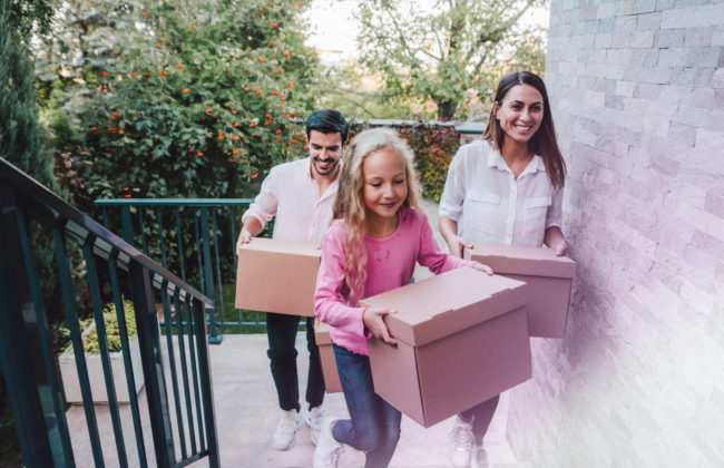 Portrait of happy family with one child moving to new house with cardboard boxes. A young family move into their new home. Smiling family carrying boxes into new home on moving day. Family entering in their new house.
