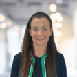 Professional head-and-shoulders portrait of a person with long dark hair, wearing a dark jacket and a green-trimmed top, positioned against a softly blurred indoor background with warm lights.