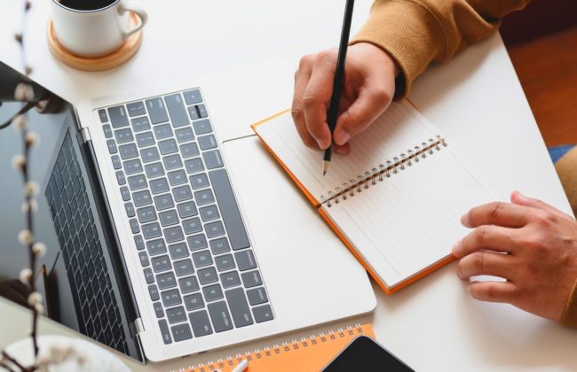 Person writes in notebook while looking at laptop