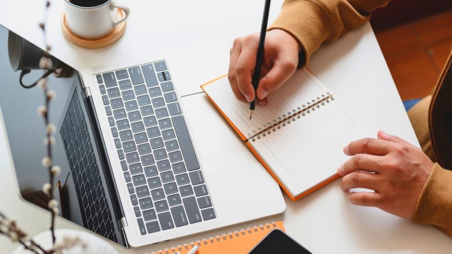 Person writes in notebook while looking at laptop