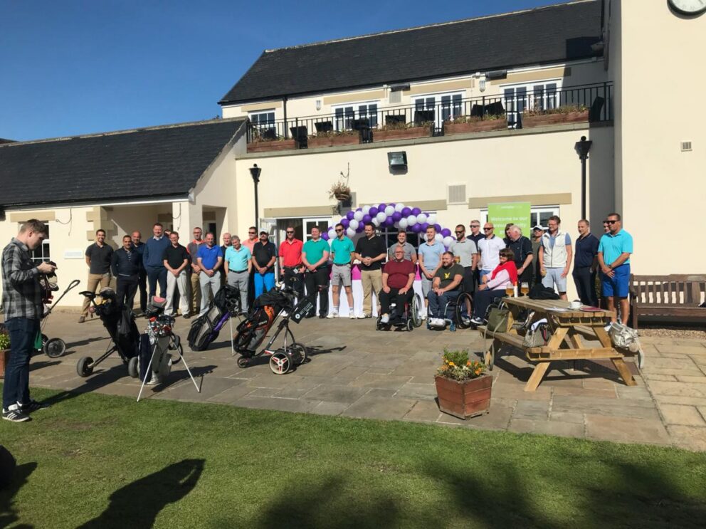 Group photo outside a light-colored building on a sunny day, with a large group of people assembled in front of an entrance decorated with a purple and white balloon arch. Several golf bags and trolleys are positioned in the foreground, suggesting a golf-related event. Some attendees use wheelchairs, while others stand or sit on benches. A picnic table with drinks and personal items is visible to the right, and a person holding a camera stands to the left capturing the scene.