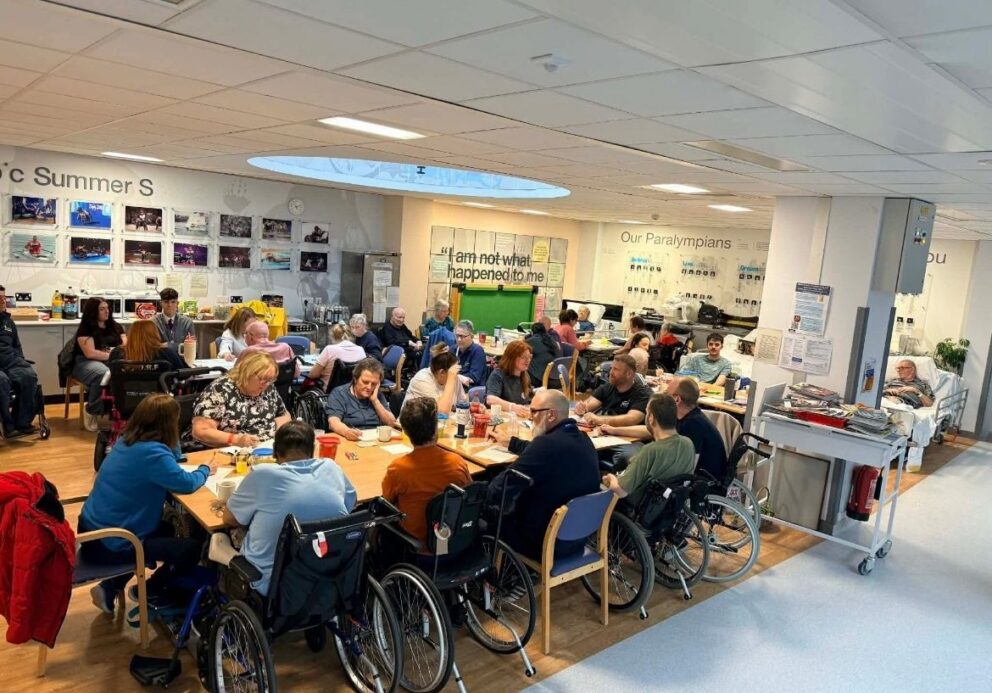Large indoor communal space with multiple tables where a group of people, including several wheelchair users, sit together engaged in a shared activity such as crafts or conversation. Drinks and materials are spread across the tables. The room has wooden flooring, bright overhead lighting, and walls displaying photos and posters, including a board titled “Our Paralympians” and a quote reading “I am not what happened to me.” In the background, a hospital-style bed and equipment are visible, indicating a rehabilitation or care setting.