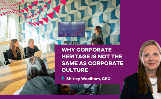 LinkedIn promotional graphic showing a meeting taking place in a modern conference room. Several colleagues are seated around a large wooden table while another colleague presents beside a wall-mounted screen displaying a presentation. The room features a geometric blue-and-white patterned wall, large windows, and pink triangular graphic accents extending across the top left corner. A purple text panel overlays the image with the headline: “WHY CORPORATE HERITAGE IS NOT THE SAME AS CORPORATE CULTURE” and the byline: “Shirley Woolham, CEO.” A professional headshot appears on the right side of the graphic against a purple background. The design uses Minster Law branding colours throughout.