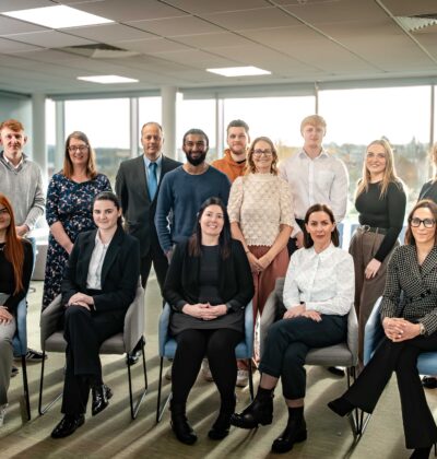 A large group team photo of Minster Law colleagues, some standing and some seated, smiling together in a bright office space with large windows overlooking the surrounding area.