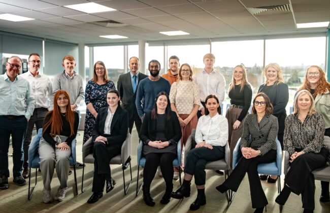A large group team photo of Minster Law colleagues, some standing and some seated, smiling together in a bright office space with large windows overlooking the surrounding area.