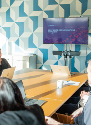 A wide view of colleagues seated around a boardroom table for a Corporate Communications - November meeting, with a presenter standing beside a screen displaying the Minster Law logo.