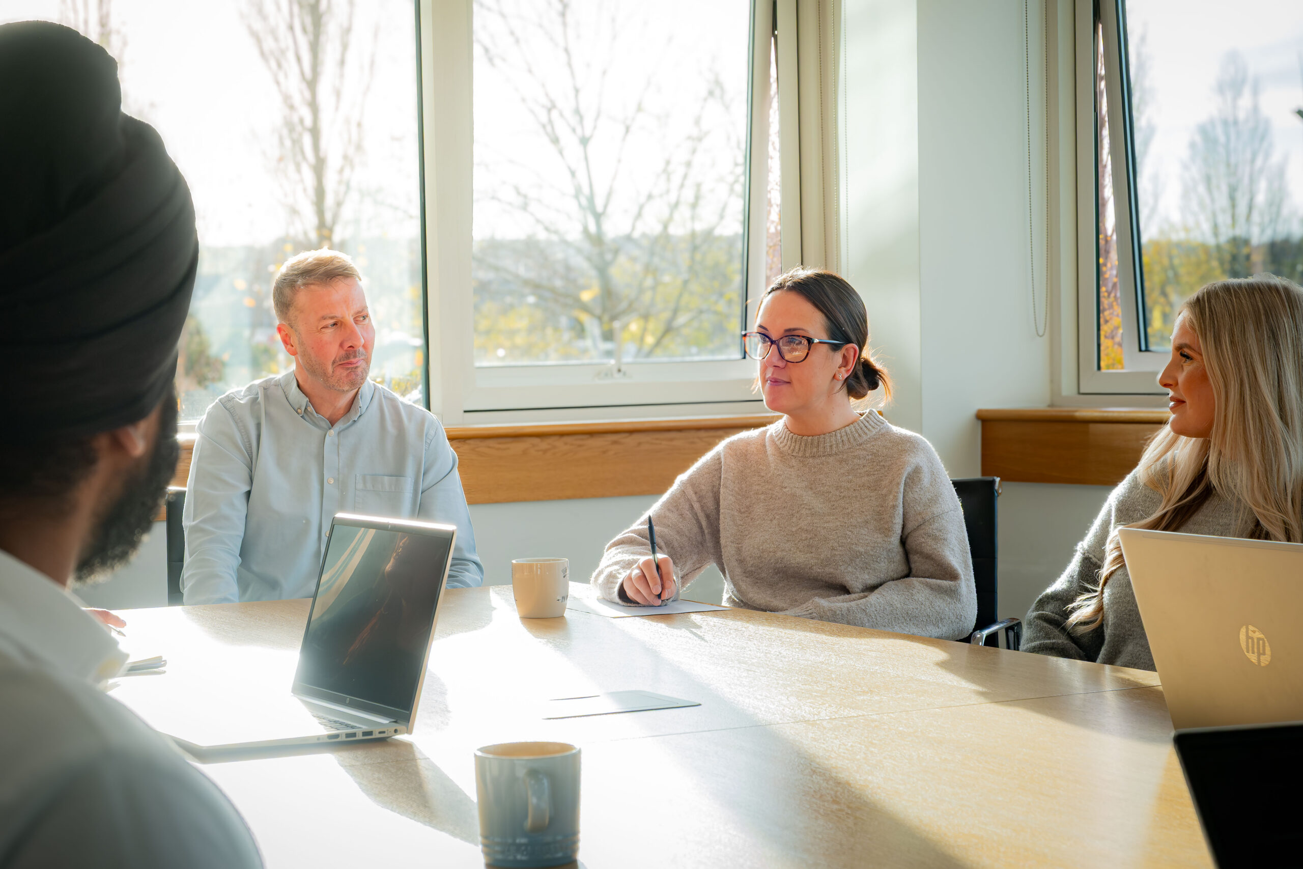 Colleagues sit around a sunlit meeting table with laptops and coffee mugs, engaged in discussion, with a woman writing notes as others look on.