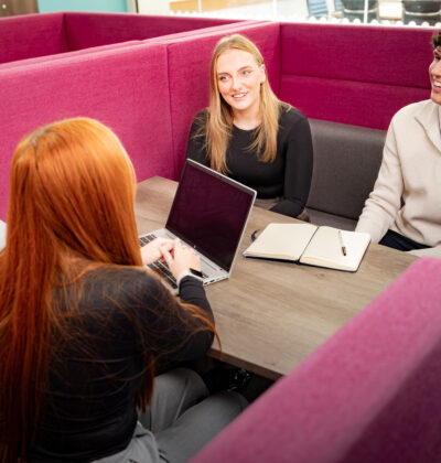 Four colleagues sit around a table in a magenta booth seating area, smiling and chatting, with a laptop and open notebook on the table between them