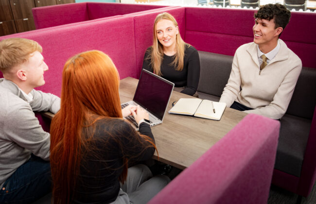 Four colleagues sit around a table in a magenta booth seating area, smiling and chatting, with a laptop and open notebook on the table between them