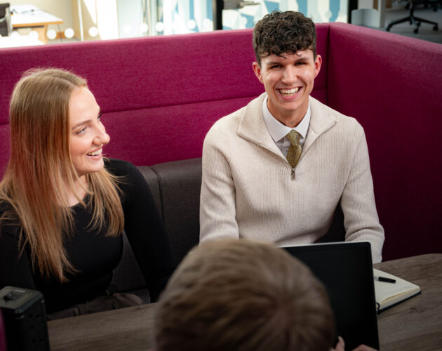 Two colleagues smile and laugh together in a magenta booth seating area, with a laptop and notepad on the table and a third colleague visible in the foreground.