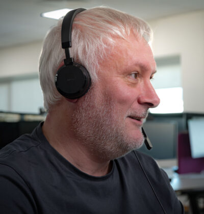Side profile of a man with white hair and a beard, wearing a Jabra headset with microphone, smiling while looking off to the side in an office setting.