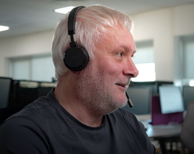 Side profile of a man with white hair and a beard, wearing a Jabra headset with microphone, smiling while looking off to the side in an office setting.
