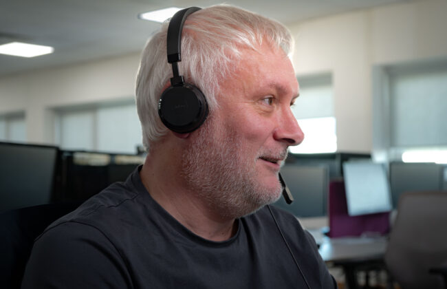 Side profile of a man with white hair and a beard, wearing a Jabra headset with microphone, smiling while looking off to the side in an office setting.