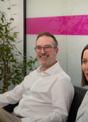 Two colleagues sit together in a meeting, a smiling man in a white shirt and a woman in a polka dot blouse, listening to a third person in the foreground, with a branded pink and white wall panel behind them.
