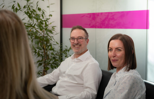 Two colleagues sit together in a meeting, a smiling man in a white shirt and a woman in a polka dot blouse, listening to a third person in the foreground, with a branded pink and white wall panel behind them.