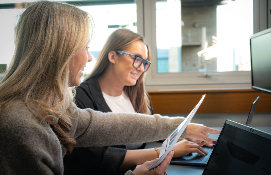Two colleagues collaborating at a desk in an office. One, with blonde hair and wearing a grey knit jumper, is smiling and holding a document with coloured charts while pointing towards a laptop screen. The other, wearing glasses and a black blazer over a white top, is smiling and typing on the laptop keyboard.