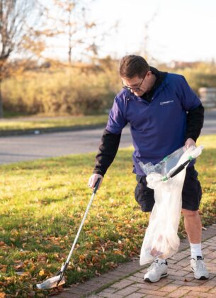 A man wearing a Minster Law polo shirt uses a litter picker to collect rubbish from grass beside a path, holding a bag for collected waste, during an outdoor community litter pick.