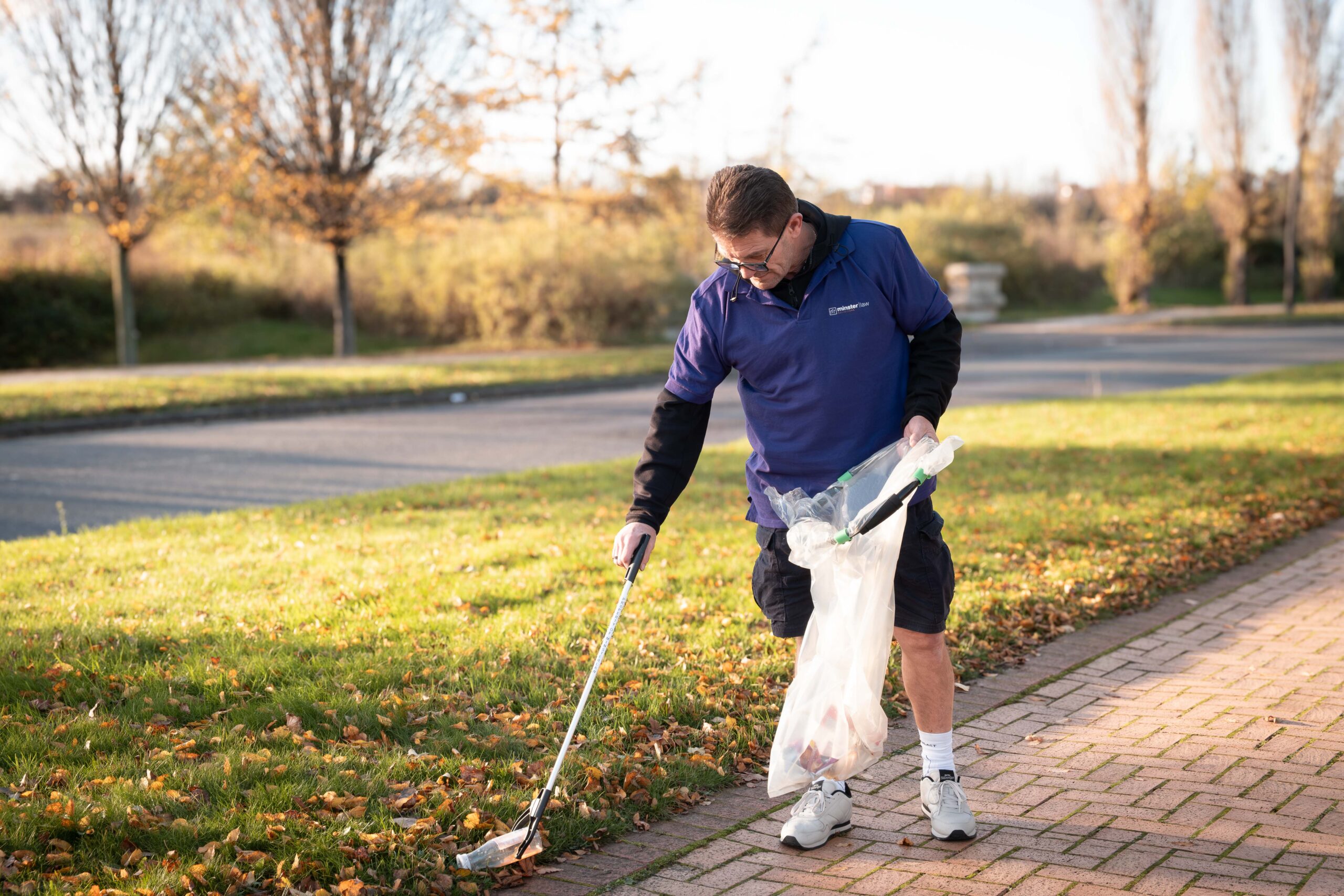 A man wearing a Minster Law polo shirt uses a litter picker to collect rubbish from grass beside a path, holding a bag for collected waste, during an outdoor community litter pick.