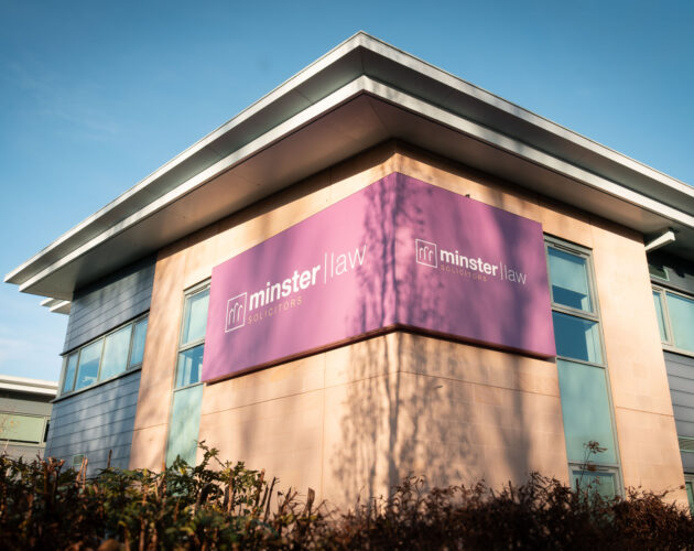 Exterior corner view of the Minster Law Solicitors office building, with the company's purple wraparound signage displayed on the building's facade against a clear blue sky.