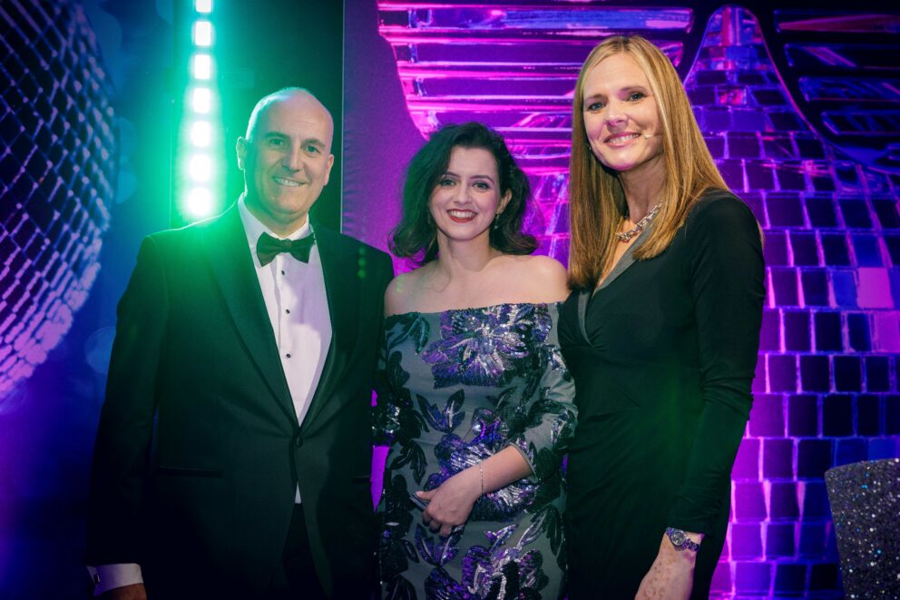 Three people dressed in formal evening wear pose together on a stage with vibrant purple, blue and green lighting and a large disco-ball themed backdrop behind them.