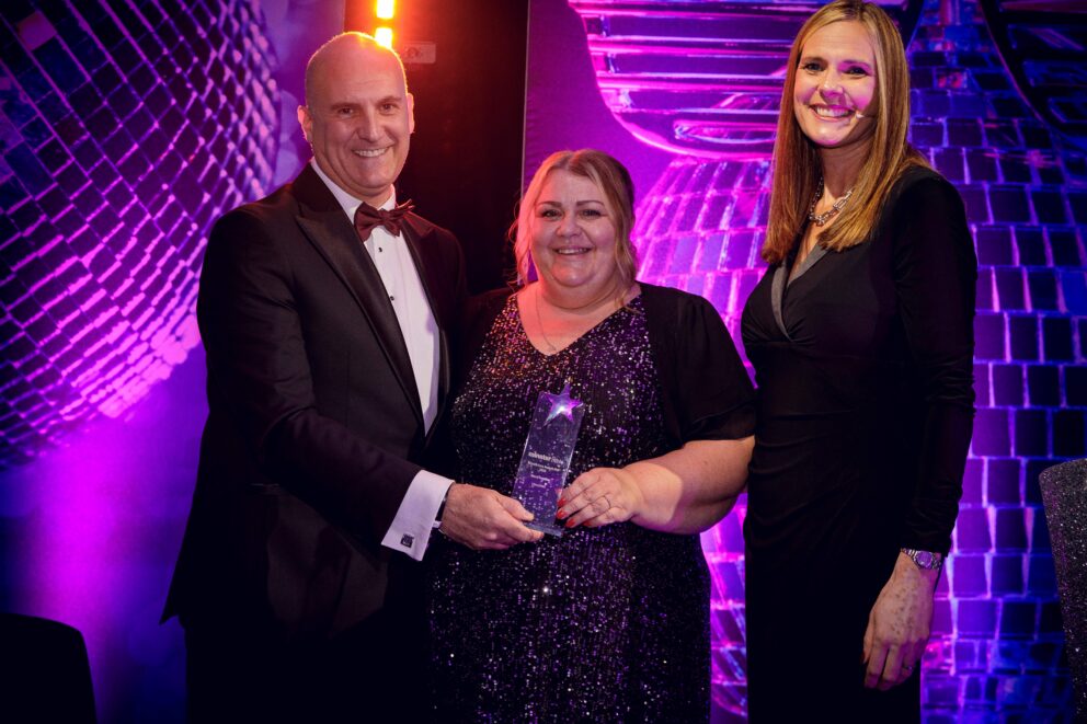 Three people stand on an awards stage under colourful lighting, with the person in the centre holding a star-shaped glass trophy. A disco-ball backdrop and glittering stage décor are visible in the background.