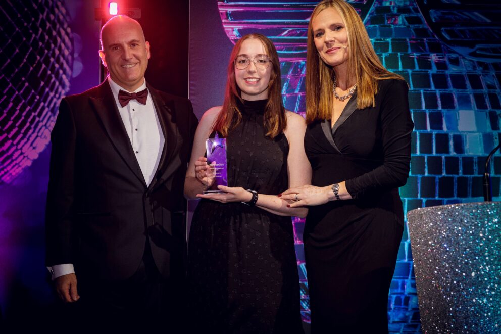 Three people pose on an awards stage with colourful purple and blue lighting. The person in the centre holds a star-shaped glass award, with a disco-ball themed backdrop and sparkling lectern visible behind them.