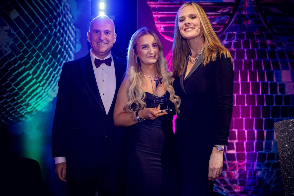 Three people in formal evening attire pose together on an awards stage. The person in the centre holds a star-shaped glass trophy, standing between two presenters. The stage is illuminated with purple, blue and pink lighting, with a large disco ball-themed backdrop visible behind them.