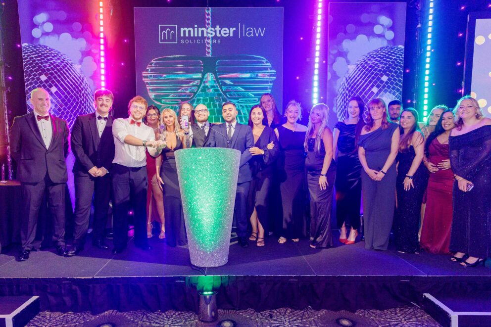 A large group of people in formal evening attire stand together on a stage behind a glittering podium at an awards ceremony. The stage is illuminated with bright purple, pink and blue lights, with large disco ball graphics and a backdrop displaying the Minster Law logo. Several group members gather around the podium while others stand along the stage, posing for a group photograph.