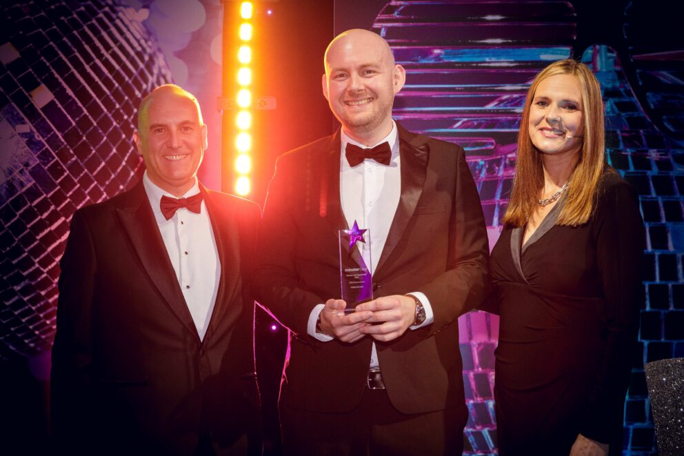 Three people in formal evening attire pose on an awards stage, with the person in the centre holding a star-shaped glass trophy. The stage is illuminated by bright pink, purple and blue lighting, with a large disco ball-themed backdrop and vertical light display visible behind them.
