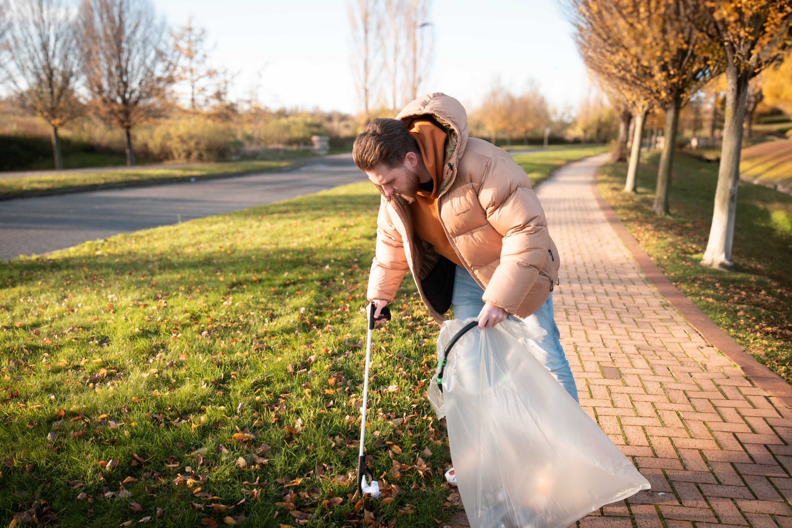 A person wearing a beige puffer jacket, light blue jeans and trainers uses a litter picker to collect rubbish from a grassy area beside a paved pathway. The person holds a large transparent refuse bag while picking up litter from the ground. The scene is set in a park or green public space with trees, grass and a roadway visible in the background. The image is taken in warm autumn sunlight, with fallen leaves scattered across the grass and path.